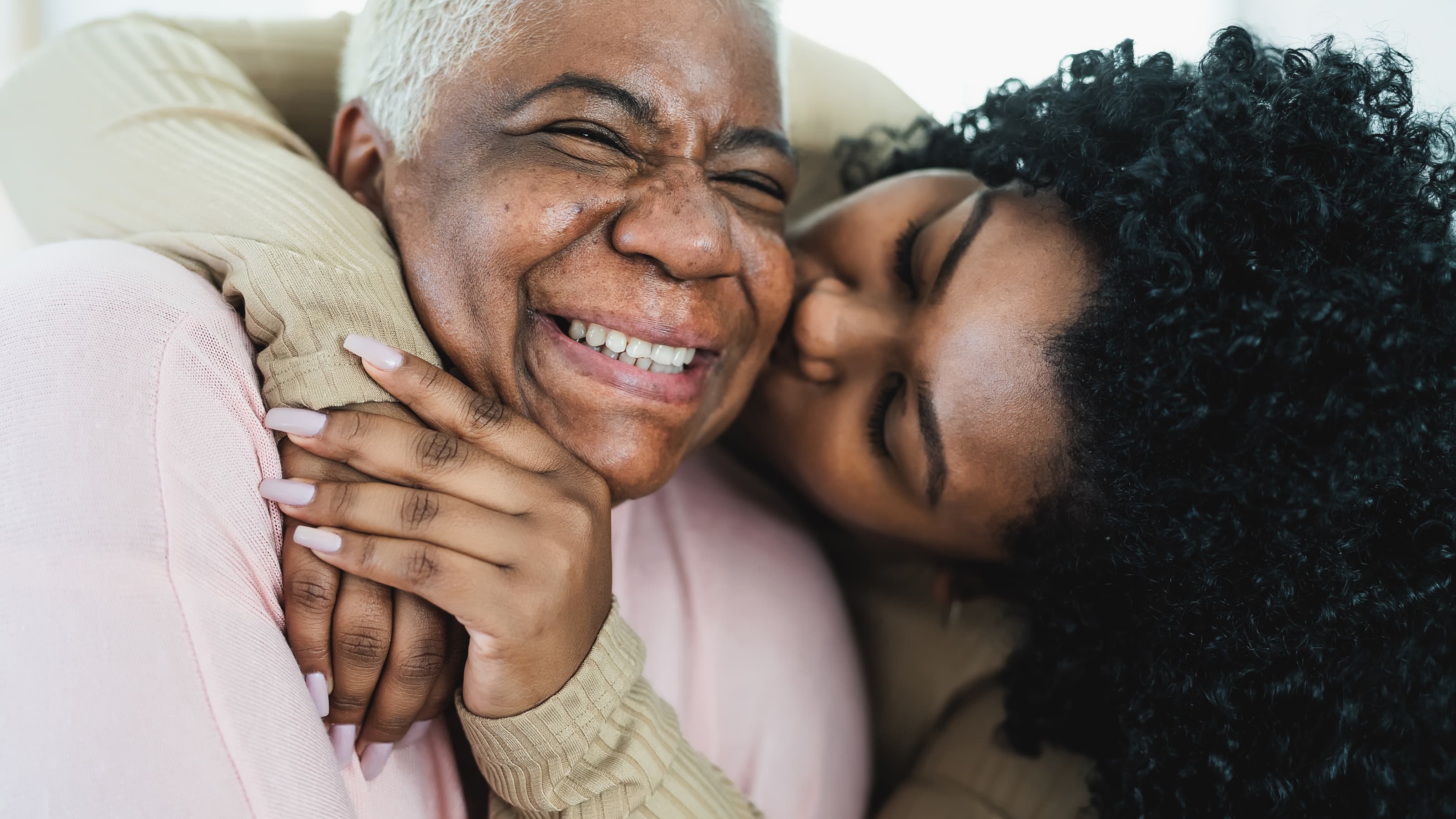 An elderly woman in a pink sweater smiles joyfully as a younger woman in beige embraces her from behind and kisses her cheek.