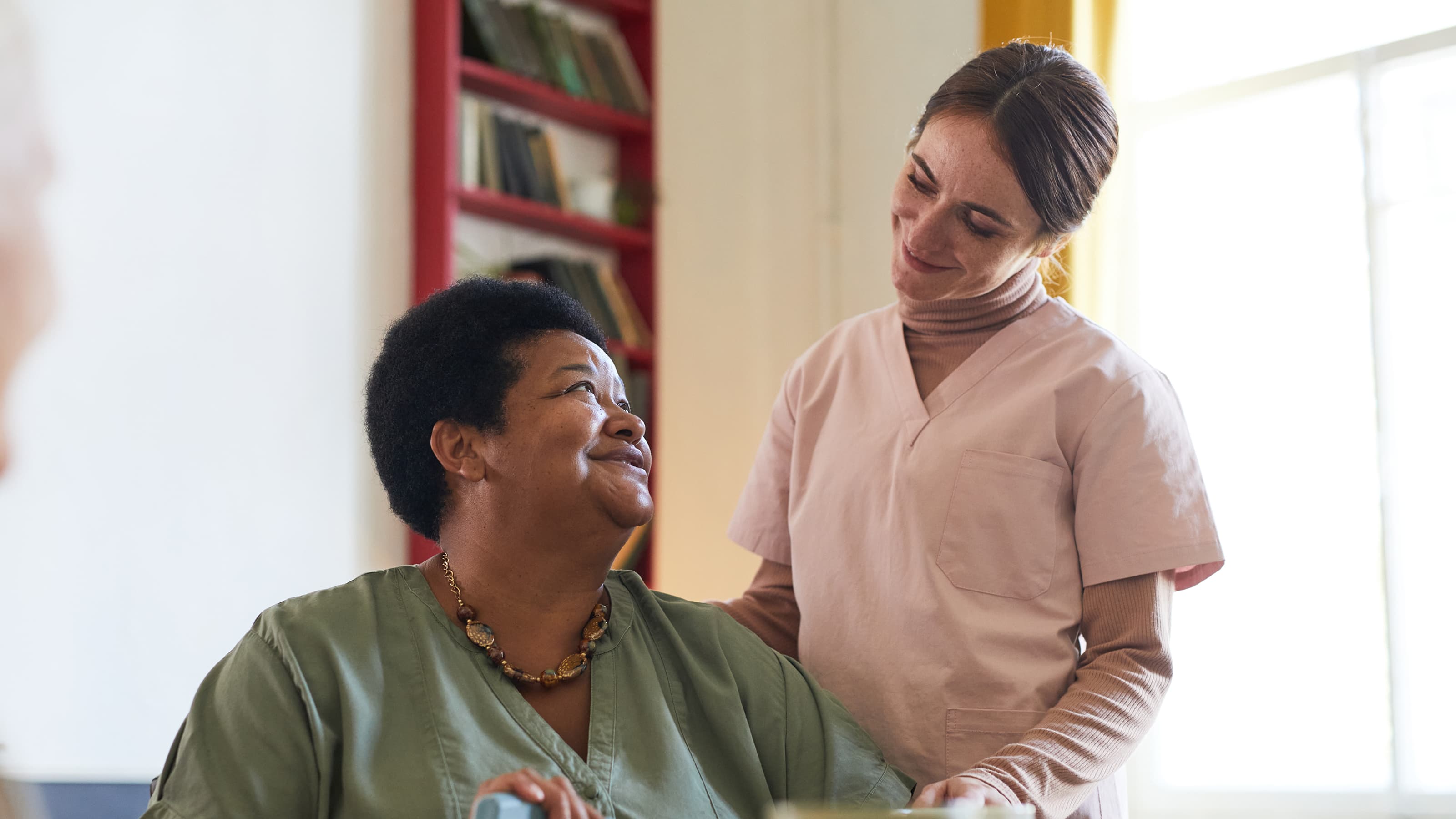 A Helper in a light pink uniform smiles warmly beside a seated woman in a green blouse and beaded necklace. The woman looks up with appreciation, surrounded by bookshelves in a bright, supportive space..